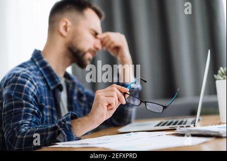 Tired male freelancer takes his glasses off and hold his hand on his face with with closed eyes, sitting at the work desk in home office, exhausted from work. He is rubbing the eyes, need a rest Stock Photo