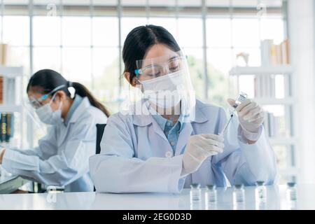 Beautiful young female scientist wearing face mask and shield performing medical research in hospital laboratory with colleague Stock Photo