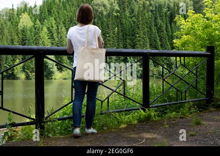 Woman carrying empty reusable shopping bag near bridge railing mockup. Female hand holding canvas eco friendly tote bag mock up Stock Photo