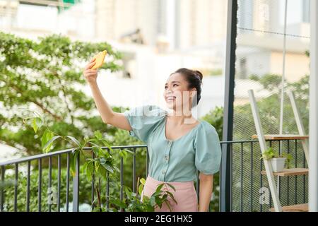 Cheerful young Asian girl having a video call with her phone when standing o balcony. Stock Photo
