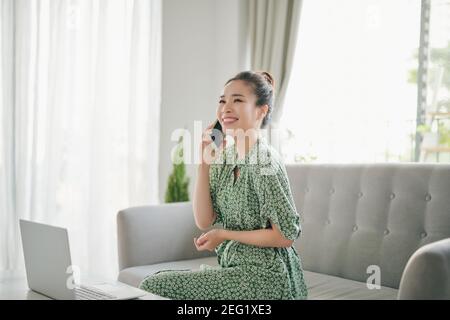 Happy young woman freelancer talking on mobile phone and using laptop, working from home during isolation, copy space. Stock Photo
