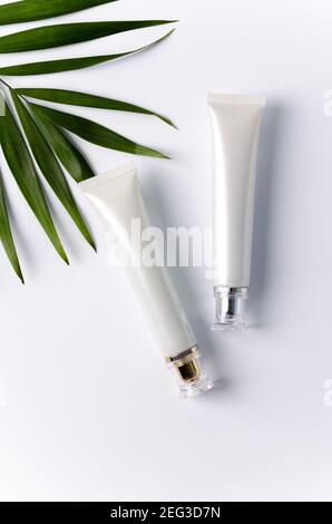 Two tubes with eye-care cream lie on a white background, next to lies a palm leaf, a vertical image, a flat layout, copying space Stock Photo