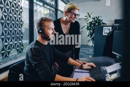 Senior manager standing by young man wearing headset working on computer. Two business professionals in call center. Stock Photo