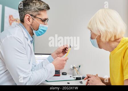 Ent doctor wearing a medical mask shows a hearing aid to a senior woman for treatment of deafness while coronavirus epidemic Stock Photo