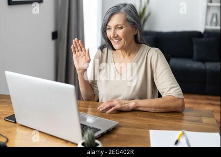 Cheerful senior woman using a laptop for video connection, remote meeting, smiling mature lady waving hello into laptop webcam, greeting online Stock Photo