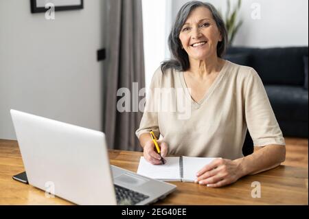 Charming mature mid-aged woman watching online webinars on the laptop sitting at the table at home, taking notes, e-learning. Senior business lady Stock Photo