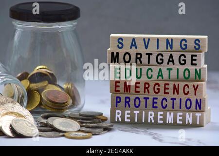 Text on wood block and glass jars with multicurrency coins - Savings, mortgage, educations, emergency, protection, retirement Stock Photo