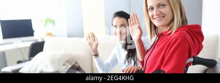 Young disabled woman in wheelchair with helper is sitting by laptop screen and waving hello Stock Photo