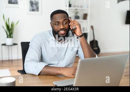 Surprised Afro American young man looking at the screen of the laptop, talking on the phone, printing, enjoing freelance work online. Front view Stock Photo