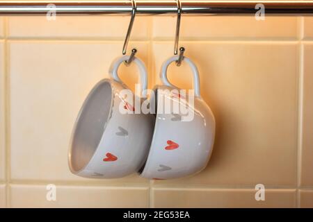 Two coffee cups with the image of hearts hanging on hooks above a table in the kitchen. Space for text. Valentine's Day Concept Stock Photo