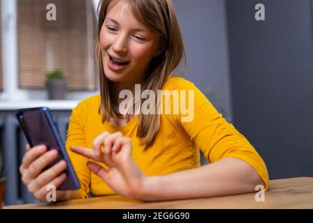 Image of happy nice, blonde, young woman in pajamas, smiling and using cellphone while sitting on the floor in living room Stock Photo