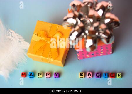 A Happy Easter inscription of multicolored beads, a yellow and pink gift boxes on a delicate blue or white background. Easter background flatly. Stock Photo