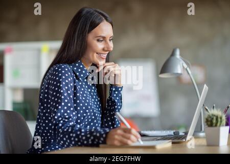 Profile side photo of young happy positive good mood smiling businesswoman writing in notebook working in laptop at office Stock Photo