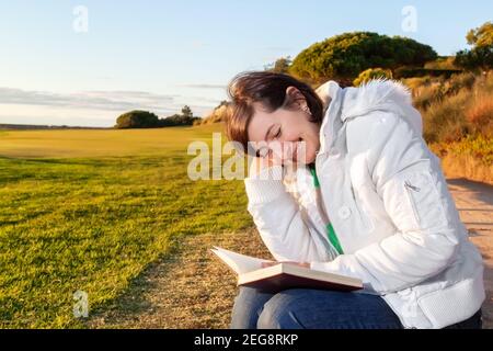 Dreamy photo of middle aged woman reading a book while sitting on a nice grassy area, during the sunset. Stock Photo