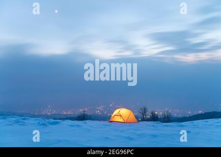 Yellow tent lighted from the inside against the backdrop of glowing city lights in fog. Amazing snowy landscape. Tourists camp in winter mountains. Travel concept Stock Photo