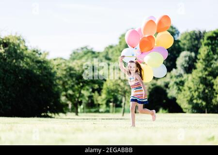 Young girl running on the grass field with colorful balloons. Childhood happiness. Stock Photo