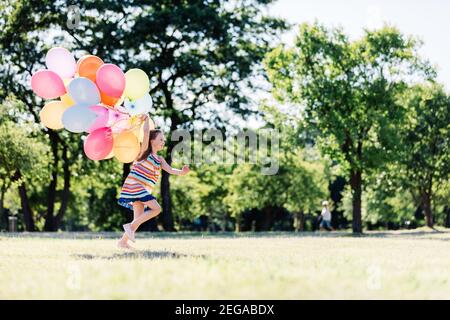 Little girl running fast with a bunch of colorful balloons in the park. Childhood activities. Stock Photo
