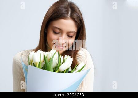 Closeup portrait of young woman holding large bouquet of white tulips on gray background. A beautiful brown-haired girl closed her eyes and breathes i Stock Photo
