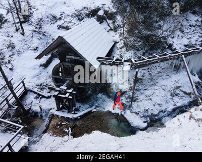 cool male Hiker with beard, with backpack, orange cap and orange pants is next to icy stream and water wheel surrounded by icicles and snow in winter Stock Photo