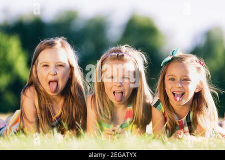 Three little girls sticking their tongues out, laying on the grass in the park. Little rebels. Sisterhood. Stock Photo