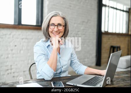 Intelligent elderly gray-haired businesswoman using a laptop in the office. Smart mature woman entrepreneur looks at the camera sitting at the desk Stock Photo