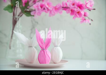 Three eggs bunny in a colored paper wrapper on a plate, nearby branch of cherry blossoms. The Easter holiday is on the table Stock Photo