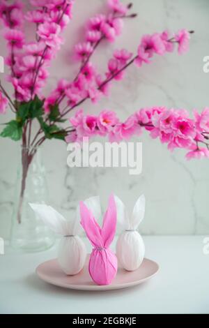 Three eggs bunny in a colored paper wrapper on a plate, nearby branch of cherry blossoms. The Easter holiday is on the table Stock Photo