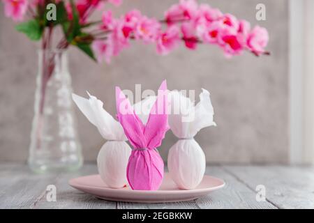 Three eggs bunny in a colored paper wrapper on a plate, nearby branch of cherry blossoms. The Easter holiday is on the table Stock Photo