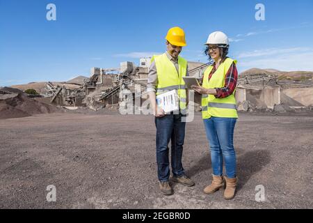 Full body of positive female engineer showing tablet to male colleague and explaining details of project while visiting industrial construction site t Stock Photo