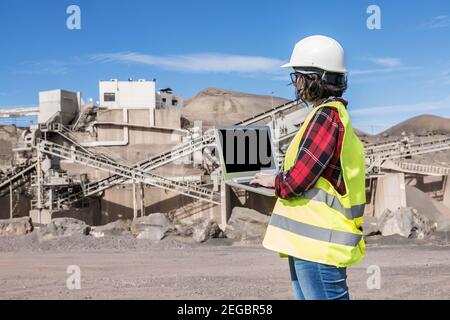 Side view of female engineer in hardhat and waistcoat working on laptop with black screen while visiting construction site of unfinished industrial fa Stock Photo