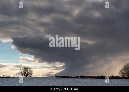 dramatic grey shower clouds with blue sky and sun rays and showing  snow falling onto snow covered fields Stock Photo