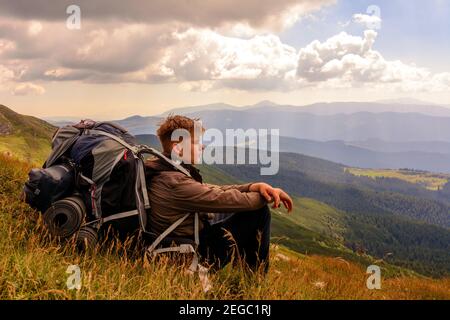 A young tourist sits on a background of mountains with a backpack, relaxing and admiring the views of the Carpathians in Ukraine.2020 Stock Photo