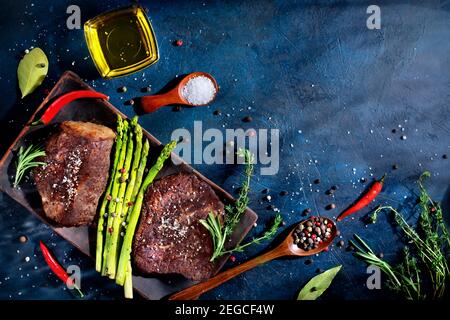 two steaks, grilled beef with vegetables, asparagus, spices, red pepper on a blue stone background. Stock Photo