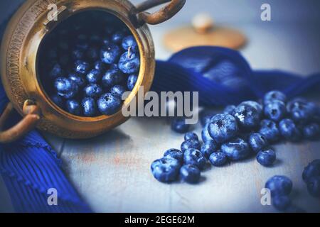 An old golden Greek bowl fell on its side on the table and spilled out the ripe blue berries of delicious blueberries. Still-life. Stock Photo