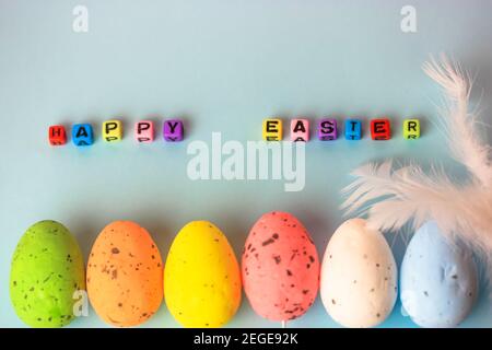 Multicolored Easter decorative eggs and white feather on a gently blue background. Happy Easter inscription made of multicolored beads. Minimalistic b Stock Photo