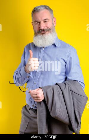Studio portrait mature bearded businessman dressed in gray suit shows thumb up, successful business concept, yellow background Stock Photo