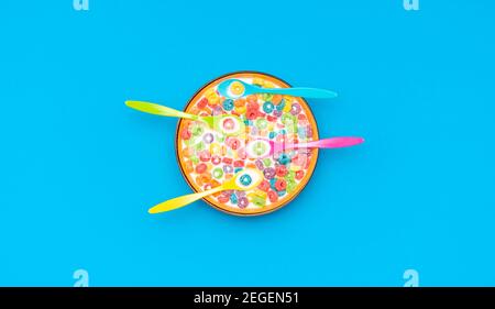 Top view with multicolored fruit rings cereals with milk in a bowl. Cereal bowl with four multicolored spoons isolated on a colored background. Stock Photo