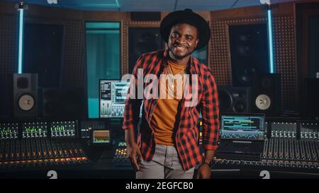 Portrait of Young Black Artist, Musician, Audio Engineer, Producer Wearing Stylish Hat and Standing in Music Record Studio. In the Background Control Stock Photo