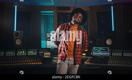 Portrait of Young Black Artist, Musician, Audio Engineer, Producer Wearing Stylish Hat and Standing in Music Record Studio. In the Background Control Stock Photo