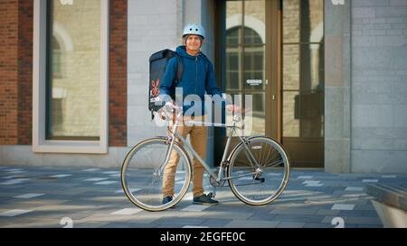 Handsome Food Delivery Man Wearing Thermal Backpack and Safety Helmet Stands Beside his Bike in the Stylish Modern City District. Portrait of the Stock Photo