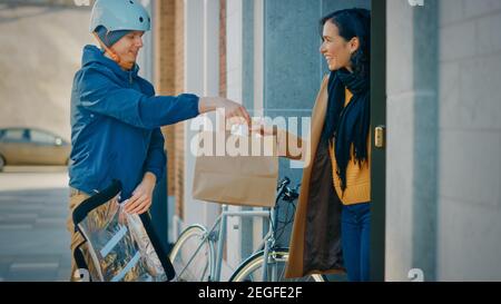 Happy Food Delivery Man Wearing Thermal Backpack on a Bike Delivers Restaurant Order to a Beautiful Female Customer. Courier Delivers Takeaway Lunch Stock Photo