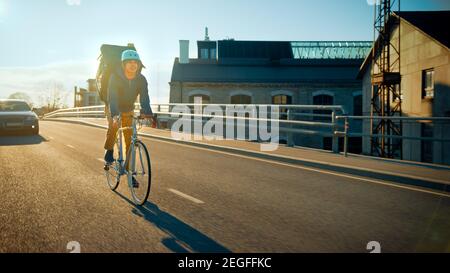 Happy Food Delivery Courier Wearing Thermal Backpack Rides a Bike on Road To Deliver Orders for Clients and Customers. Sunny Day in Modern City with Stock Photo