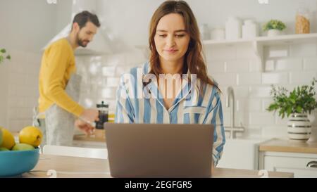 Young Beautiful Woman Using Laptop Computer While Wearing Blue Pyjamas. Brunette Female Sitting in a Modern Sunny Kitchen Room. Freelancer Working Stock Photo
