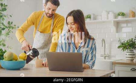 Young Beautiful Woman Working on Laptop Computer Wearing Blue Pyjamas. Husband Pouring Cup of Hot Coffee and Encouraging His Girlfriend. Female Stock Photo