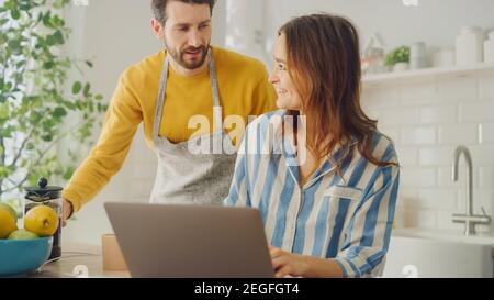 Beautiful Young Couple Using Laptop Computer in Kitchen at Home. Female Wearing Pyjamas. Man Wearing an Apron is Helping Girlfriend with Online Work Stock Photo