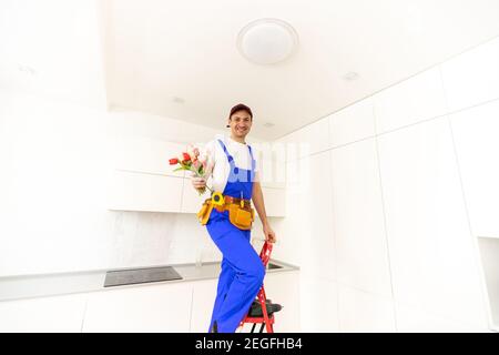 smiling handsome plumber holding bouquet flowers and looking at camera in kitchen Stock Photo