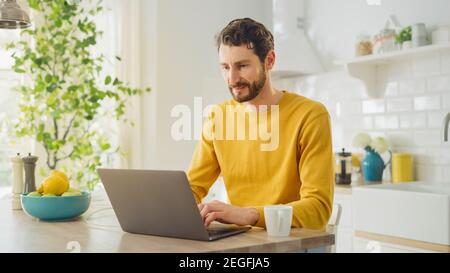 Young Handsome Man Using Laptop Computer and Drinking Morning Cup of Coffee or Tea While Wearing a Yellow Pullover. Curly-hared Male Sitting in a Stock Photo