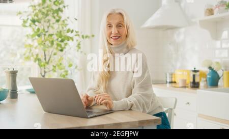 Beautiful Senior Woman Using Laptop Computer in a Sunny Kitchen. She Looks at Camera and Smiles. Grandmother is Connected with Children via internet Stock Photo
