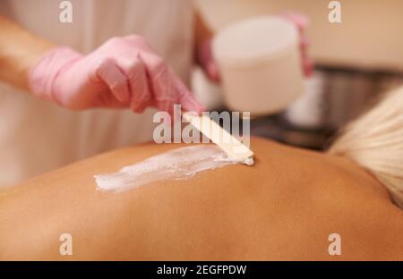 Female masseur applies a massage cream scrub using a wooden spatula to the back of a young woman lying on a massage table and enjoying a therapy sessi Stock Photo