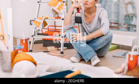 Pretty Woman Sits with Paint Brush in a Room Prepared for Renovation and Selects a Color for Painting the Walls. Repairs in the New Apartment. Stock Photo